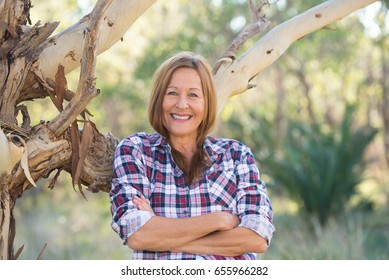 Portrait Attractive Mature Woman In Rural Country, Wearing Plaid Shirt, Posing Relaxed With Happy Smile Against Australian Outback Bush Background.