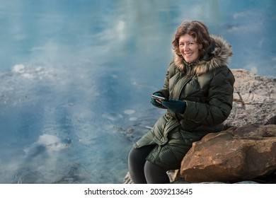 Portrait Of Attractive Mature Woman Sitting On Large Stone By Pond In Midwestern Park With Her Smartphone; Winter In Missouri, Midwest