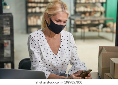 Portrait of attractive mature woman, small business owner wearing protective mask using smartphone, receiving orders while sitting at the desk in her craft pottery shop - Powered by Shutterstock