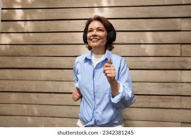 Portrait of attractive mature woman with wireless headphones against background of siding cottage wall. Middle aged brunette in a blue shirt listening to the music. Active lifestyle for the elderly.