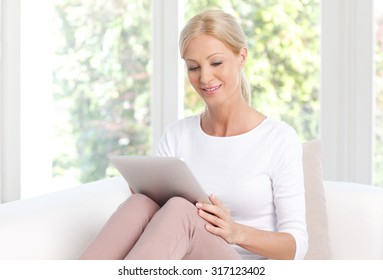 Portrait Of Attractive Middle Age Woman Holding In Her Hands Digital Tablet While Sitting At Living Room And Working Online.