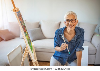 Portrait attractive middle aged professional female painter and gray hair working at home standing in front easel  Happy mature woman painting   watching at camera  