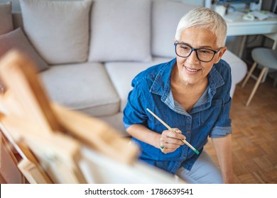 Portrait of attractive middle aged professional female painter with gray hair working at home standing in front of easel, having thoughtful look, finishing still life painting - Powered by Shutterstock