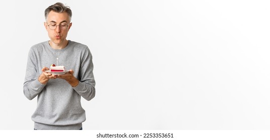 Portrait of attractive middle-aged man in grey sweater and glasses, celebrating his birthday, blowing a candle on b-day cake and making wish, standing over white background. - Powered by Shutterstock