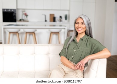 Portrait of attractive middle-aged woman with long silver hair sitting in modern apartment, mid-aged multiracial asian female resting at home, looking at camera and smiling with wide toothy smile - Powered by Shutterstock