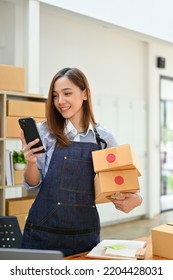 Portrait, Attractive millennial Asian female small online business owner holding shipping boxes and using her phone to read a message from customers. - Shutterstock ID 2204428031