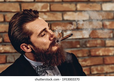 Portrait Of Attractive Pretentious Man With A Mustache And Beard Smoking A Brown Cigar Against A Brick Wall Background. Hobby Concept