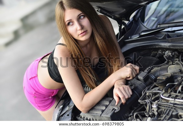 Portrait of an\
attractive and pretty young caucasian girl in pink shorts and black\
shirt with wrenches near a black car with an open hood. The concept\
of repair and maintenance of a\
car