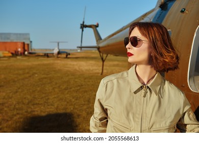 Portrait Of An Attractive Professional Woman Pilot With Bright Red Lips And Black Sunglasses Posing Next To Her Helicopter.