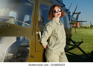 Portrait Of An Attractive Professional Woman Pilot With Bright Red Lips And Black Sunglasses Standing By A Helicopter.