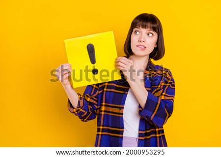 Portrait of attractive puzzled girl holding exclamation sign board biting lip isolated over bright yellow color background