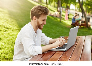 Portrait of attractive red-head man with beard in white shirt, sitting in the park, laughing and doing his job on laptop. Freelance concept.