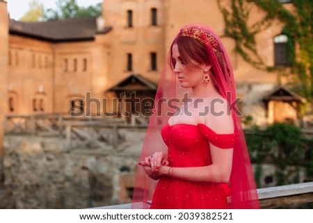Portrait of attractive redhead tattooed woman in long red dress, diadema and red veil on blurred medieval castle background