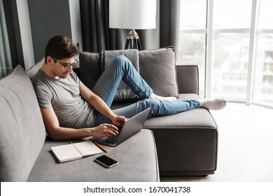 Portrait of an attractive serious young bearded man wearing casual clothes sitting on a couch at the living room, using laptop computer