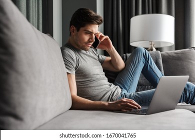 Portrait Of An Attractive Serious Young Bearded Man Wearing Casual Clothes Sitting On A Couch At The Living Room, Talking On Mobile Phone While Using Laptop Computer