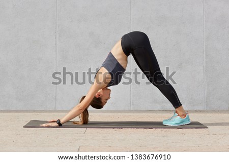 Portrait of attractive slim sporty woman in yoga pose at sportstudio, dressed sportwears, has pony tail, poses against grey background. Well shaped young girl trains indoor alone. Health care concept.