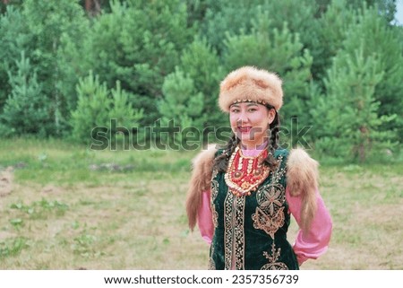 Portrait of attractive smiling Asian girl in fur headdress and traditional festive Bashkir clothes, looking at camera, at rural holiday.