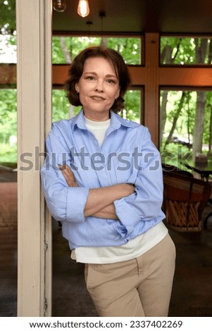 Portrait of an attractive smiling elderly woman. Mature caucasian woman with crossed hands. Middle-aged brunette in casual clothes on the porch of a country cottage. Active lifestyle for the elderly.