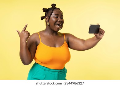 Portrait of attractive smiling Nigerian woman with stylish hairstyle holding mobile phone recording video isolated on yellow background. Beautiful female plus size blogger using smartphone - Powered by Shutterstock