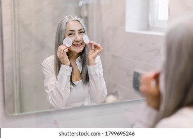 Portrait Of Attractive Smiling Senior Woman With Natural Gray Hair, Cleaning Her Face With Cotton Pads, Caring For Her Skin In A Bathroom, Looking In The Mirror. Back View, Reflection In The Mirror