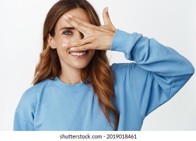 Portrait of attractive smiling woman open eye, peeking through fingers and looking happy, sneak peak at smth, standing in blue shirt against white background - Shutterstock ID 2019018461