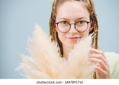 Portrait of an attractive smiling woman in round glasses with stylish blond afro braids over blue background. Holding fluffy cereal bouquet.