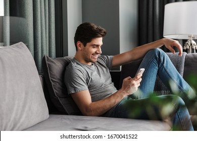 Portrait of an attractive smiling young bearded man wearing casual clothes sitting on a couch at the living room, using mobile phone