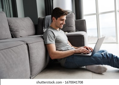 Portrait of an attractive smiling young bearded man wearing casual clothes sitting on a floor at the couch in the living room, using laptop computer