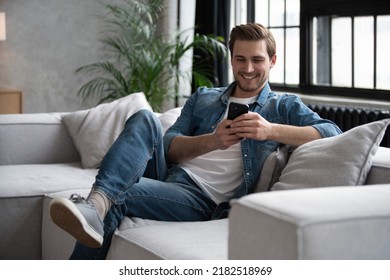 Portrait of an attractive smiling young man wearing casual clothes sitting on a couch at the living room, using mobile phone - Powered by Shutterstock