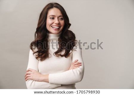 Portrait of an attractive smiling young woman with long brunette hair wearing sweater standing isolated over gray background, arms folded