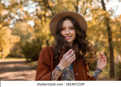portrait of attractive stylish smiling woman with long curly hair walking in park dressed in warm brown coat autumn trendy fashion, street style wearing hat