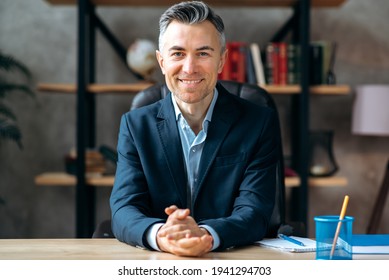 Portrait of an attractive, successful, influential, adult caucasian manager, businessman or lawyer in stylish formal suit, sitting at work desk in the office, looks at the camera friendly smiling