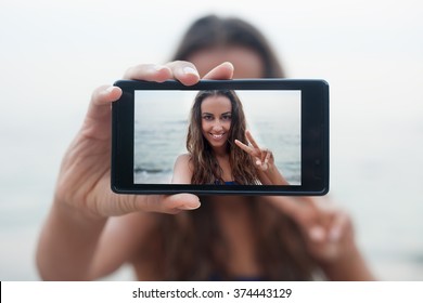 Portrait of attractive teenager girl standing on a summer sandy beach on holiday, holding a smartphone device taking selfies pictures of herself on vacation against blue sky. People travel technology. - Powered by Shutterstock