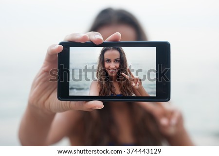 Portrait of attractive teenager girl standing on a summer sandy beach on holiday, holding a smartphone device taking selfies pictures of herself on vacation against blue sky. People travel technology.