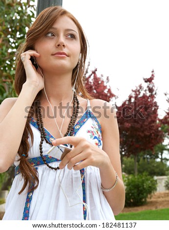 Portrait of an attractive teenager girl visiting a park during the summer and holding a smartphone with headphones, listening to music and enjoying a day out. Technology and lifestyle.