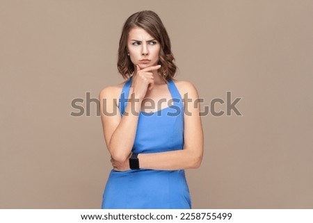 Portrait of attractive thoughtful puzzled woman with wavy hair holding chin, looking away with pensive expression, planning, wearing blue dress. Indoor studio shot isolated on light brown background.