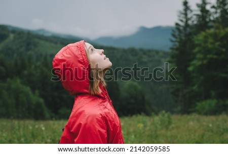 Portrait of attractive tourist girl in red raincoat stands on valley in mountains and looks up at cloudy sky. Pretty woman in raincoat hiking looking up at rain clouds.