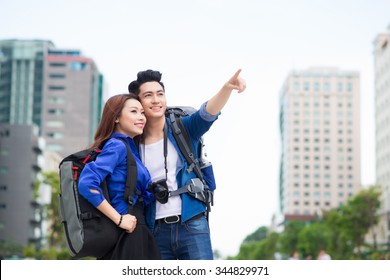 Portrait Of An Attractive Tourist Young Couple Relaxing Sightseeing And Visiting A Destination City On Holiday, Pointing Up And Enjoying Traveling Together, Outdoors. Tourism, Travel And Lifestyle.