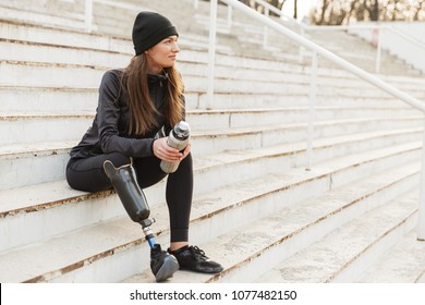 Portrait of attractive woman in black tracksuit with prosthetic leg sitting at the street stairs and holding thermos cup - Powered by Shutterstock