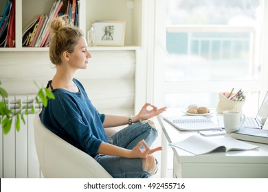 Portrait of an attractive woman in a chair at the table with cup and laptop, book, pencils, notebook on it. Lotus pose, concept photo - Shutterstock ID 492455716