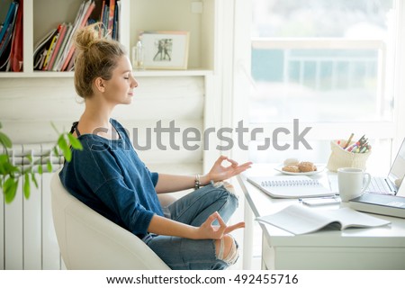 Portrait of an attractive woman in a chair at the table with cup and laptop, book, pencils, notebook on it. Lotus pose, concept photo