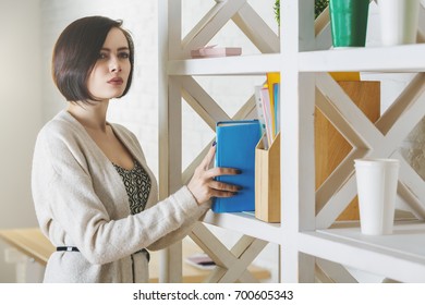 Portrait Of Attractive Woman Holding Closed Book In Modern Office Interior. Knowledge And Information Concept 