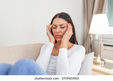 Portrait of an attractive woman sitting on a sofa at home with a headache, feeling pain and with an expression of being unwell. Upset depressed woman lying on couch feeling strong headache migraine.