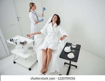 Portrait of attractive woman in white bathrobe lying with closed eyes during medical procedure. Physician checking IV infusion