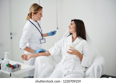 Portrait of attractive woman in white bathrobe sitting in armchair and holding glass of lemon water while physician in sterile gloves checking IV infusion