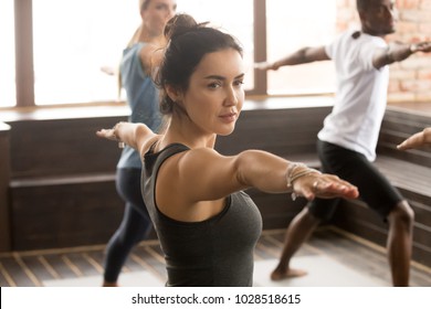 Portrait of attractive yogi girl and a group of young sporty people practicing yoga lesson, standing in Warrior two exercise, Virabhadrasana 2 pose, working out, indoor close up, studio - Powered by Shutterstock