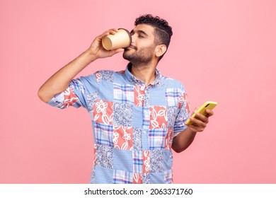 Portrait of attractive young adult man in casual style shirt standing and holding disposable cup and phone , drinking and enjoying hot beverage. Indoor studio shot isolated on pink background.