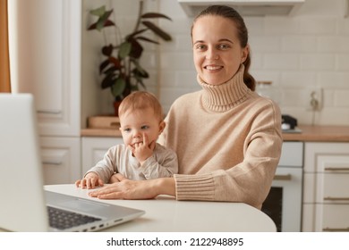 Portrait Of Attractive Young Adult Woman Wearing Beige Sweater Sitting In Kitchen And Working Online On Laptop, Holding Baby, Looking At Camera With Charming Smile.