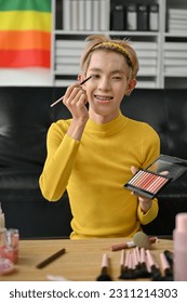 A portrait an attractive young Asian gay beauty blogger influencer is applying eyeshadow to his eyelid while recording his makeup tutorial video at home 
