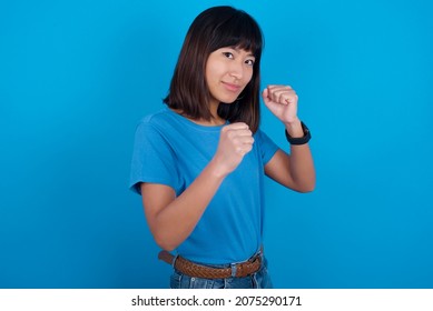 Portrait of attractive young asian woman wearing blue t-shirt against blue background holding hands in front of him in boxing position going to fight.